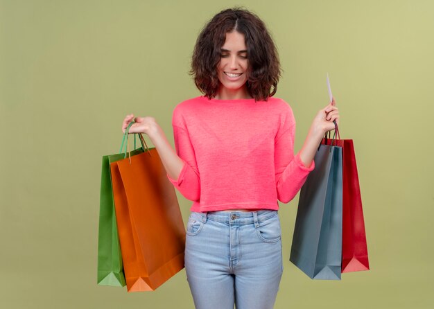 Smiling young beautiful woman holding carton bags and card on isolated green wall