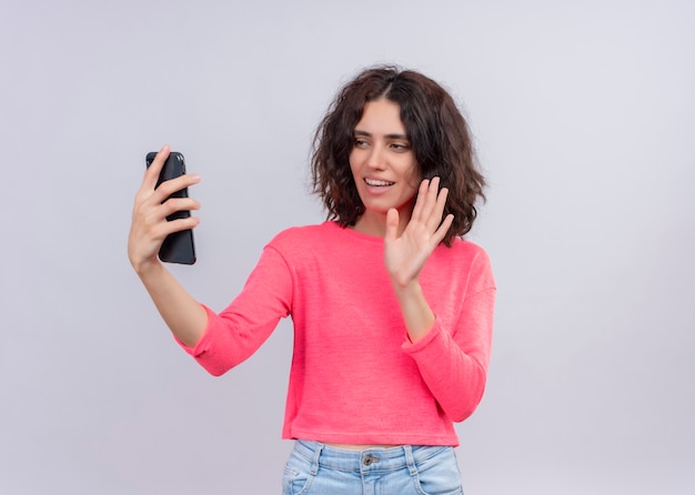 Sorridente giovane bella donna che tiene il telefono cellulare e agitando su di esso sulla parete bianca isolata