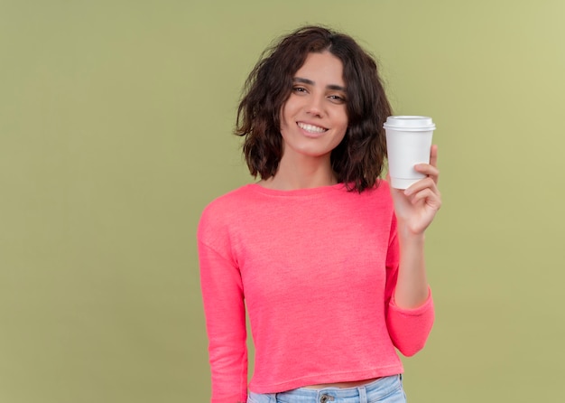 Smiling young beautiful woman holding plastic coffee cup on isolated green wall with copy space