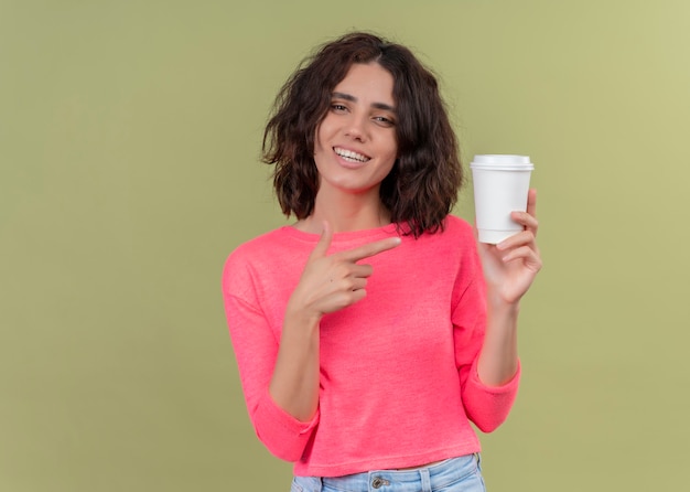 Free photo smiling young beautiful woman holding plastic coffee cup and pointing at it on isolated green wall with copy space