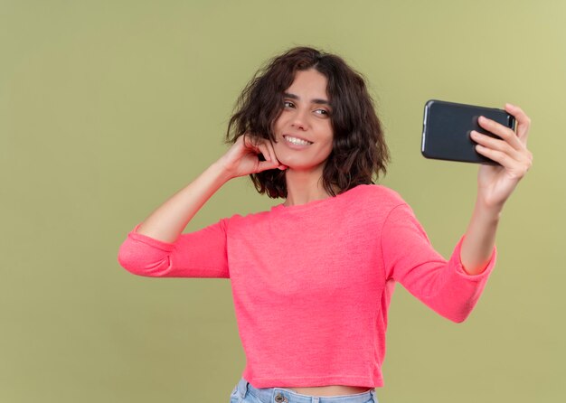 Smiling young beautiful woman putting hand on chin and taking selfie with mobile phone on isolated green wall