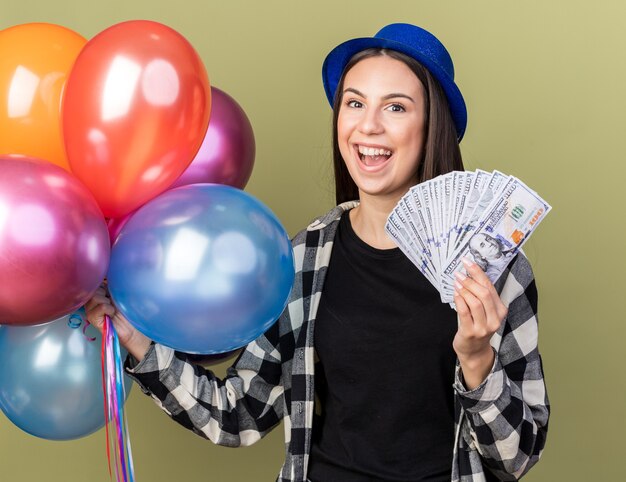 オリーブグリーンの壁に分離された現金で風船を保持している青い帽子をかぶって笑顔の若い美しい女性
