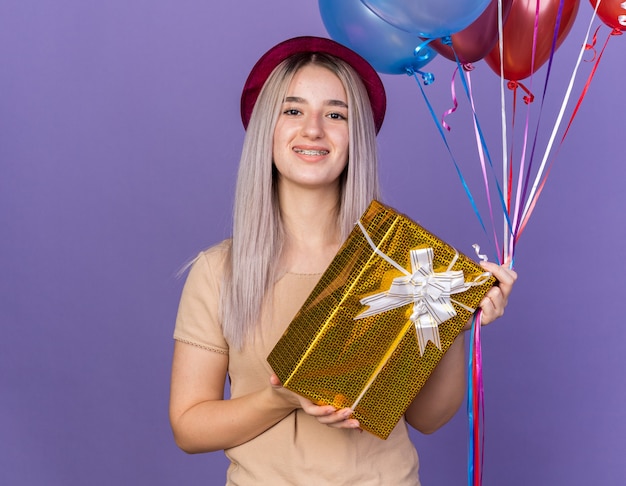 Smiling young beautiful woman wearing dental braces with party hat holding balloons with gift box isolated on blue wall