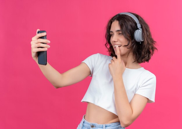 Улыбающаяся молодая красивая женщина в наушниках и держащая мобильный телефон положив палец на губу на розовой стене