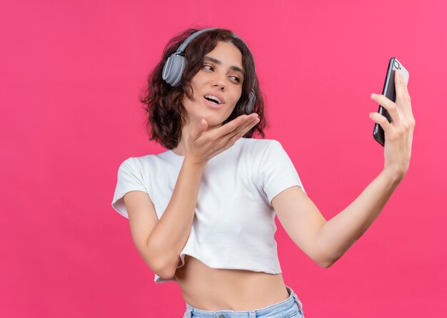 Smiling young beautiful woman wearing headphones and holding mobile phone sending blow kiss on pink wall with copy space