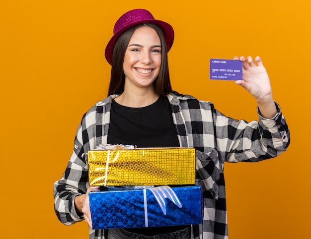 Sorridente giovane bella donna che indossa cappello da festa in possesso di scatole regalo e carta di credito isolata sulla parete arancione