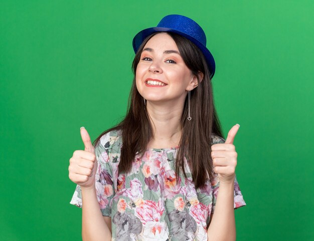 녹색 벽에 고립 된 엄지손가락을 보여주는 파티 모자를 쓰고 웃는 젊은 아름 다운 여자