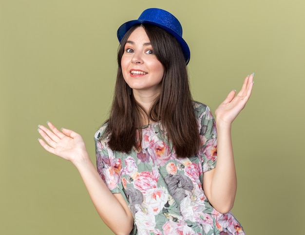 Foto gratuita sorridente giovane bella donna che indossa cappello da festa allargando le mani isolate sul muro verde oliva