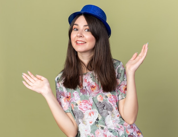 Smiling young beautiful woman wearing party hat spreading hands isolated on olive green wall