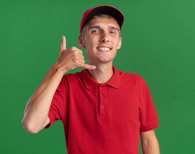 Smiling young blonde delivery boy gestures hang loose sign isolated on green wall with copy space