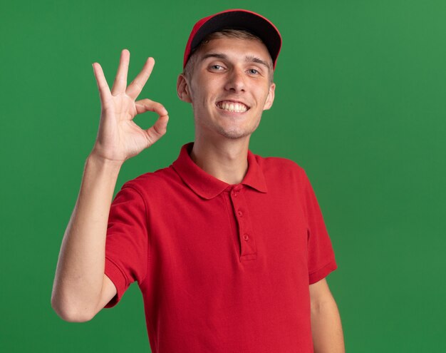 Smiling young blonde delivery boy gestures ok hand sign isolated on green wall with copy space