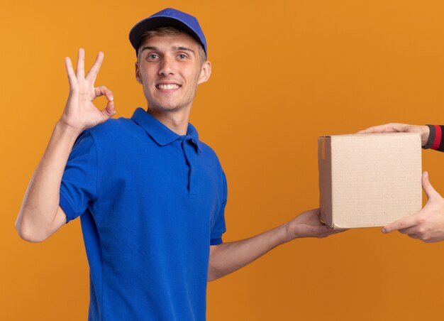 Smiling young blonde delivery boy giving cardbox to someone and gesturing ok hand sign