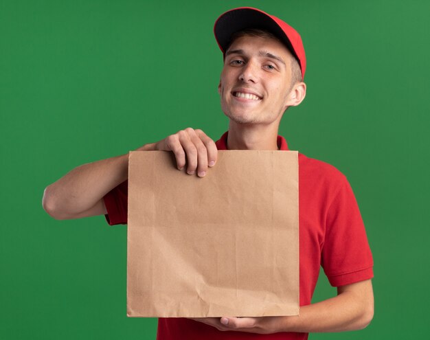 Smiling young blonde delivery boy holding paper package isolated on green wall with copy space