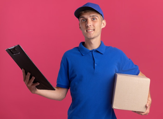 Улыбающийся молодой блондин посыльный держит картонную коробку и буфер обмена, изолированные на розовой стене с копией пространства