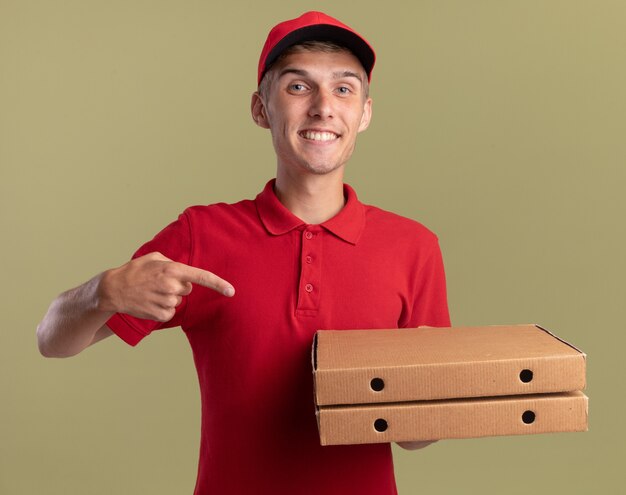 Smiling young blonde delivery boy holds and points at pizza boxes