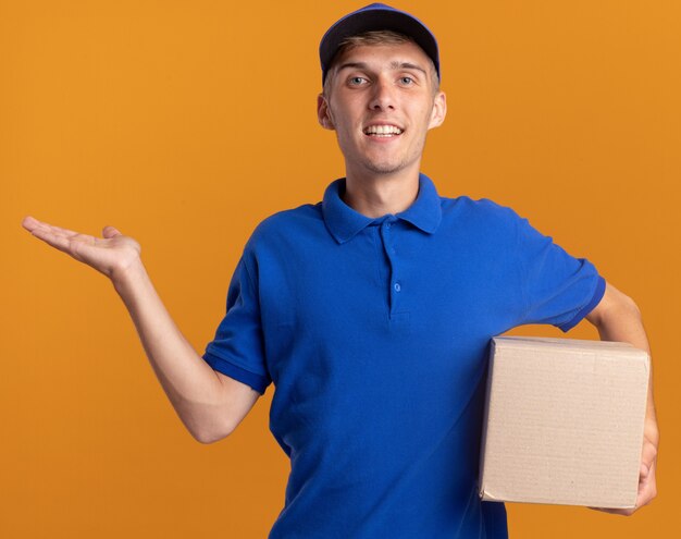 Smiling young blonde delivery boy keeps hand open and holds cardbox isolated on orange wall with copy space