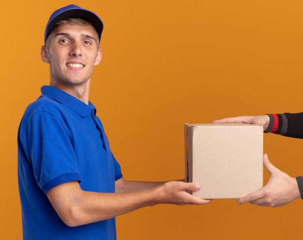 Smiling young blonde delivery boy stands sideways and gives cardbox to someone looking at camera