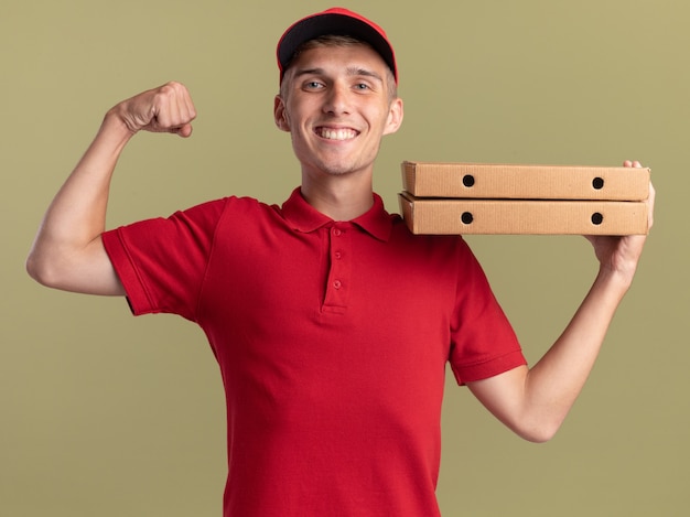 Улыбающийся молодой блондин посыльный напрягает бицепсы и держит коробки с пиццей на плече на оливково-зеленом