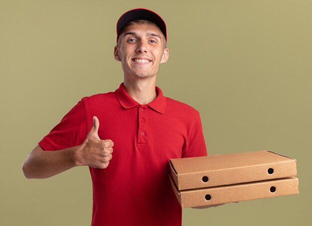 Smiling young blonde delivery boy thumbing up and holding pizza boxes on olive green 