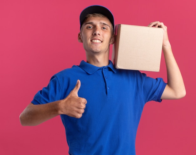 Улыбающийся молодой блондин посыльный показывает палец вверх и держит картонную коробку на плече на розовом