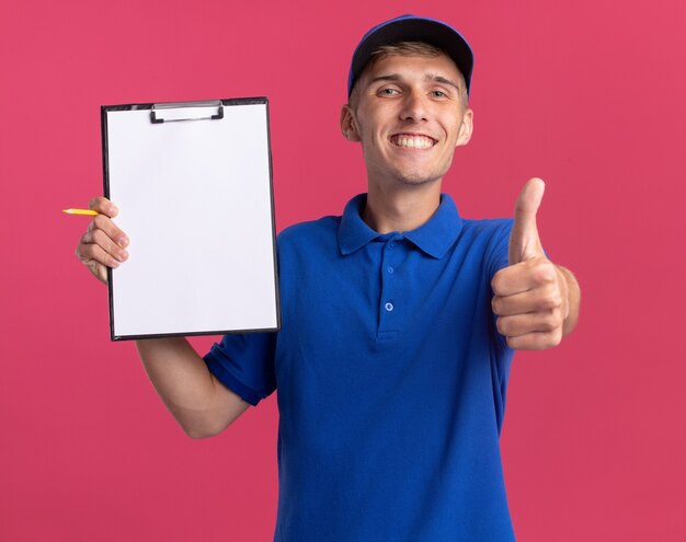 Smiling young blonde delivery boy thumbs up and holds clipboard isolated on pink wall with copy space