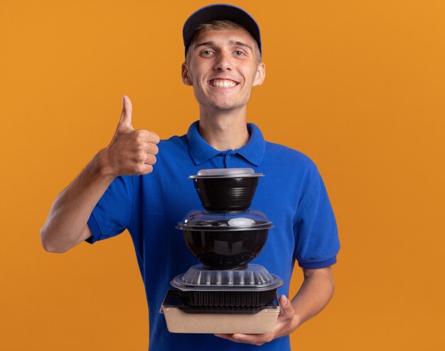 Il giovane ragazzo biondo sorridente delle consegne sfoglia in su e tiene i contenitori per alimenti sull'arancia