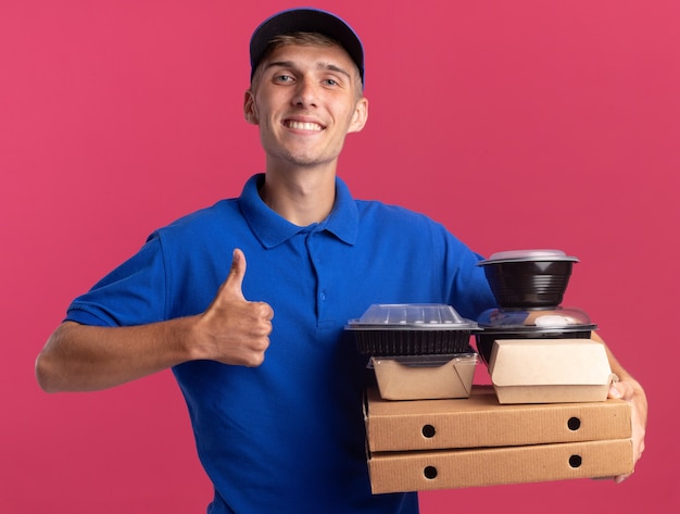 Free photo smiling young blonde delivery boy thumbs up and holds food containers and packages on pizza boxes