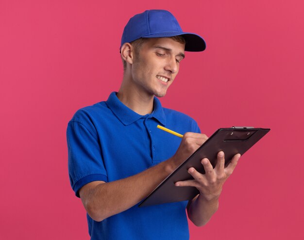 Smiling young blonde delivery boy writes on clipboard with pen isolated on pink wall with copy space