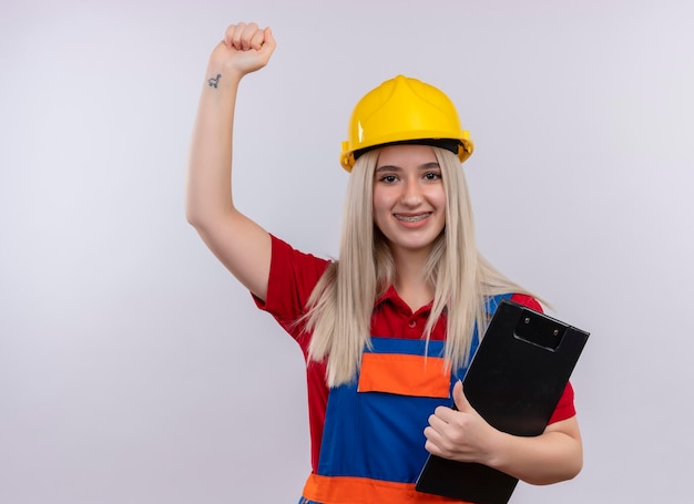 Улыбающаяся молодая блондинка инженер-строитель девушка в униформе в стоматологических скобах, держащая буфер обмена, поднимая кулак на изолированном белом пространстве