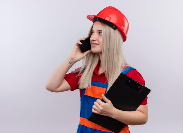 Smiling young blonde engineer builder girl in uniform in dental braces holding clipboard and talking on phone looking at left side on isolated white space with copy space