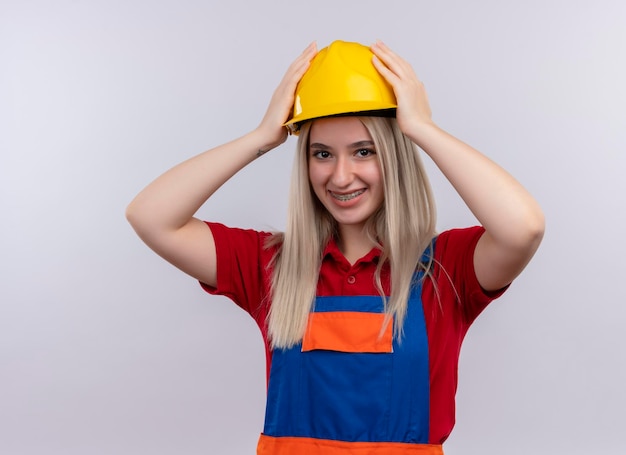 Sorridente giovane ragazza bionda ingegnere costruttore in uniforme in parentesi graffe dentali mettendo le mani sul casco di sicurezza su uno spazio bianco isolato