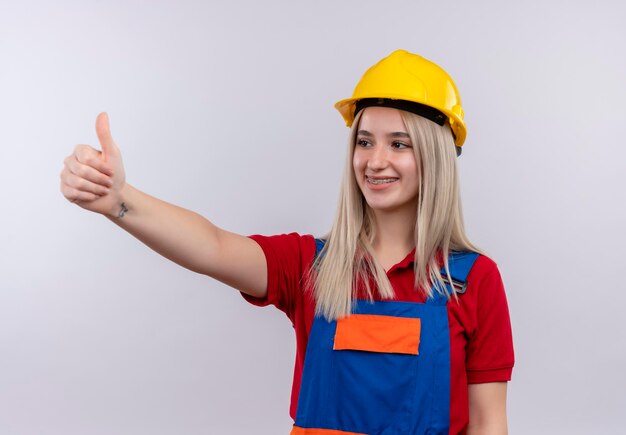 Улыбающаяся молодая белокурая девушка-строитель в униформе и зубных скобах показывает палец вверх слева на изолированном белом пространстве