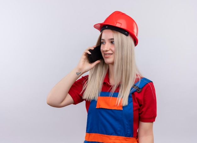 Smiling young blonde engineer builder girl in uniform in dental braces talking on phone looking at left side on isolated white space with copy space