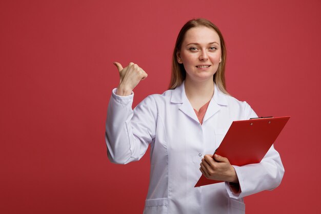 Smiling young blonde female doctor wearing medical robe and stethoscope around neck holding clipboard pointing to side 