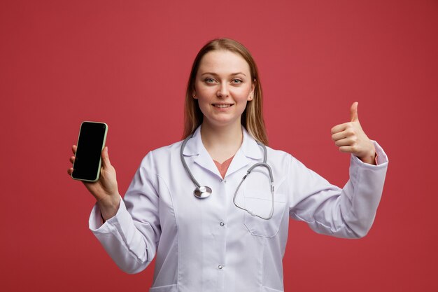 親指を上げて携帯電話を保持している首の周りに医療ローブと聴診器を身に着けている若い金髪の女性医師の笑顔