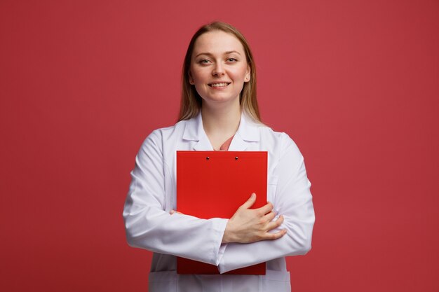 Smiling young blonde female doctor wearing medical robe and stethoscope around neck hugging clipboard 