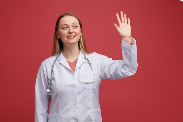 Улыбающаяся молодая блондинка женщина-врач в медицинском халате и стетоскопе на шее, глядя на размахивая рукой
