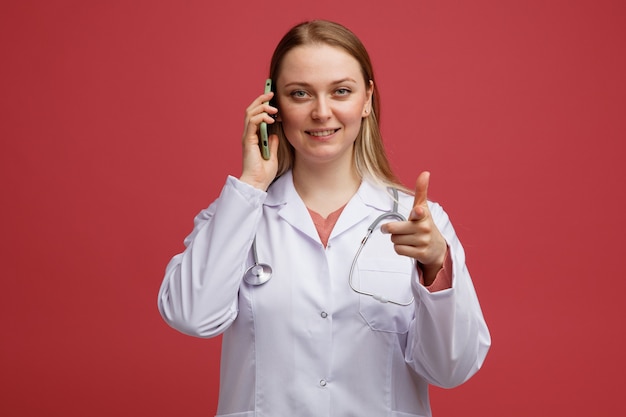 Sorridente giovane bionda dottoressa indossa veste medica e stetoscopio intorno al collo parlando al telefono guardando e indicando la fotocamera