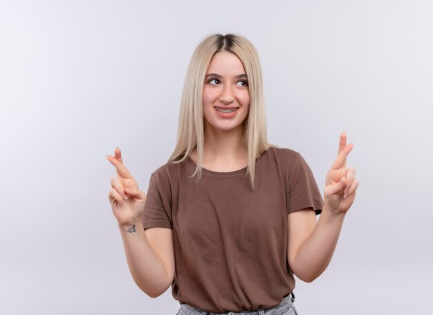 Smiling young blonde girl in dental braces doing crossed fingers gesture looking at right side on isolated white space