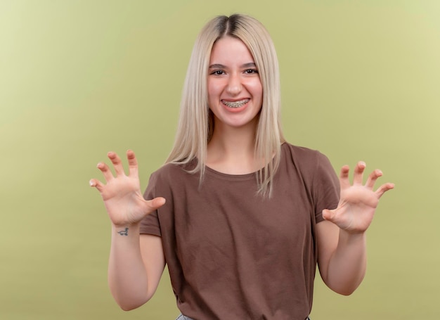 Smiling young blonde girl in dental braces doing tiger paws gesture on isolated green space