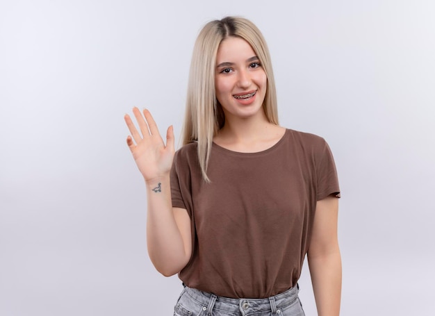 Smiling young blonde girl in dental braces gesturing hi on isolated white space with copy space