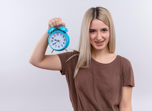 Smiling young blonde girl in dental braces holding alarm clock on isolated white space