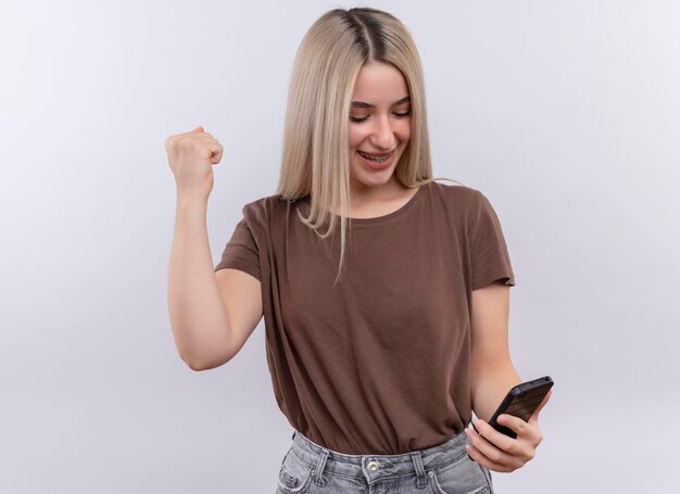 Smiling young blonde girl in dental braces holding mobile phone looking at it with raised fist on isolated white space