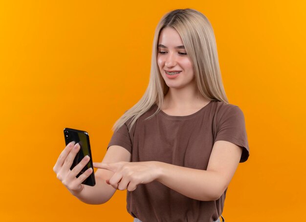 Smiling young blonde girl in dental braces holding mobile phone and touching it with finger on isolated orange space
