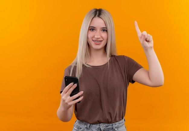 Smiling young blonde girl in dental braces holding mobile phone with raised finger on isolated orange space with copy space