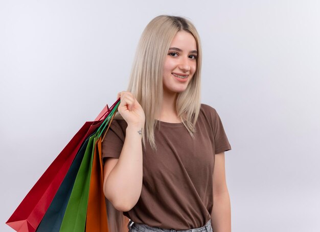 Smiling young blonde girl in dental braces holding shopping bags on shoulder on isolated white space with copy space