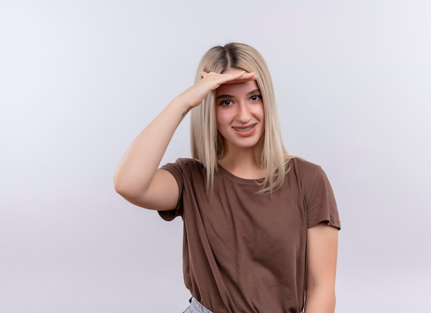 Smiling young blonde girl in dental braces looking at distance with hand on forehead on isolated white space with copy space