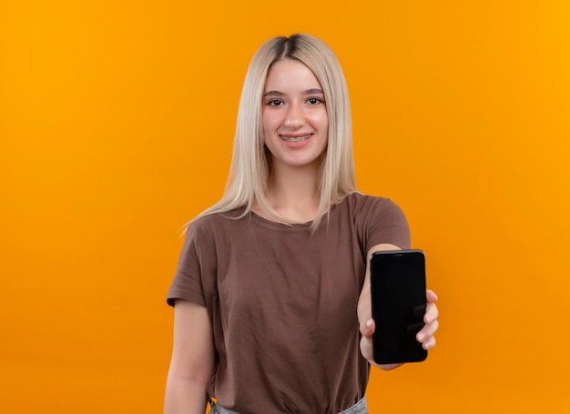 Smiling young blonde girl in dental braces stretching out mobile phone on isolated orange space with copy space