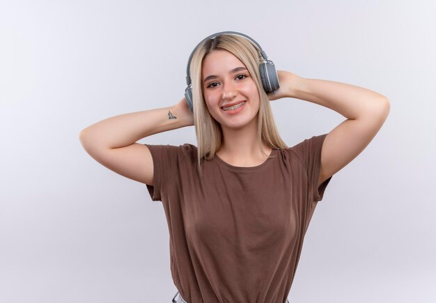 Free photo smiling young blonde girl in dental braces wearing headphones with hands behind head on isolated white space