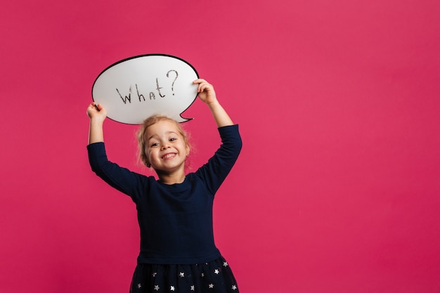 Smiling young blonde girl holding speech bubble and looking at the camera over pink wall